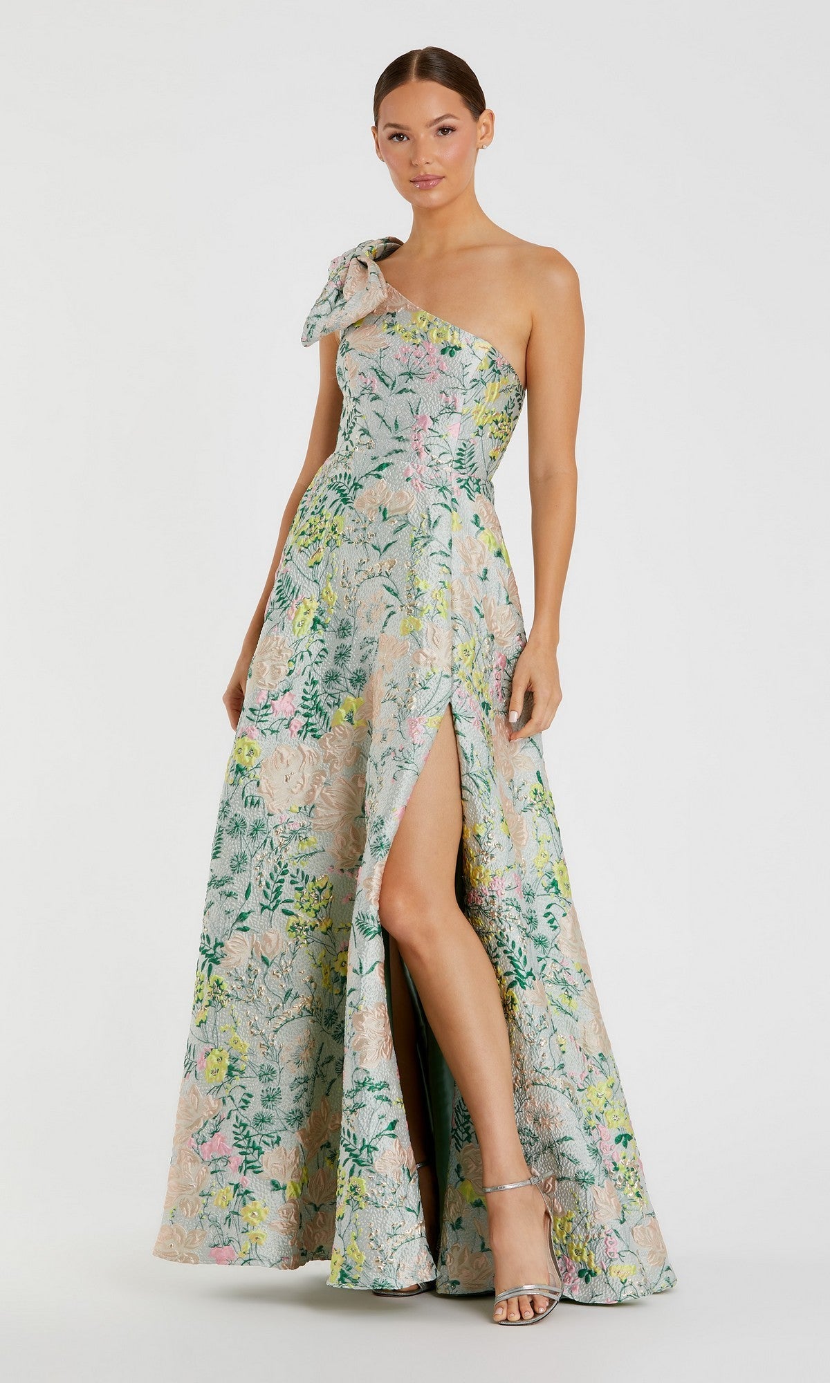 A woman models the Mac Duggal 11919 long formal dress in pastel multi—a one-shoulder, floor-length floral brocade gown with an a-line skirt and high slit—standing against a plain white background.